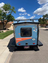 A light blue Cricket camper, a great option for an RV rental, is parked on a residential street, adorned with various travel stickers.