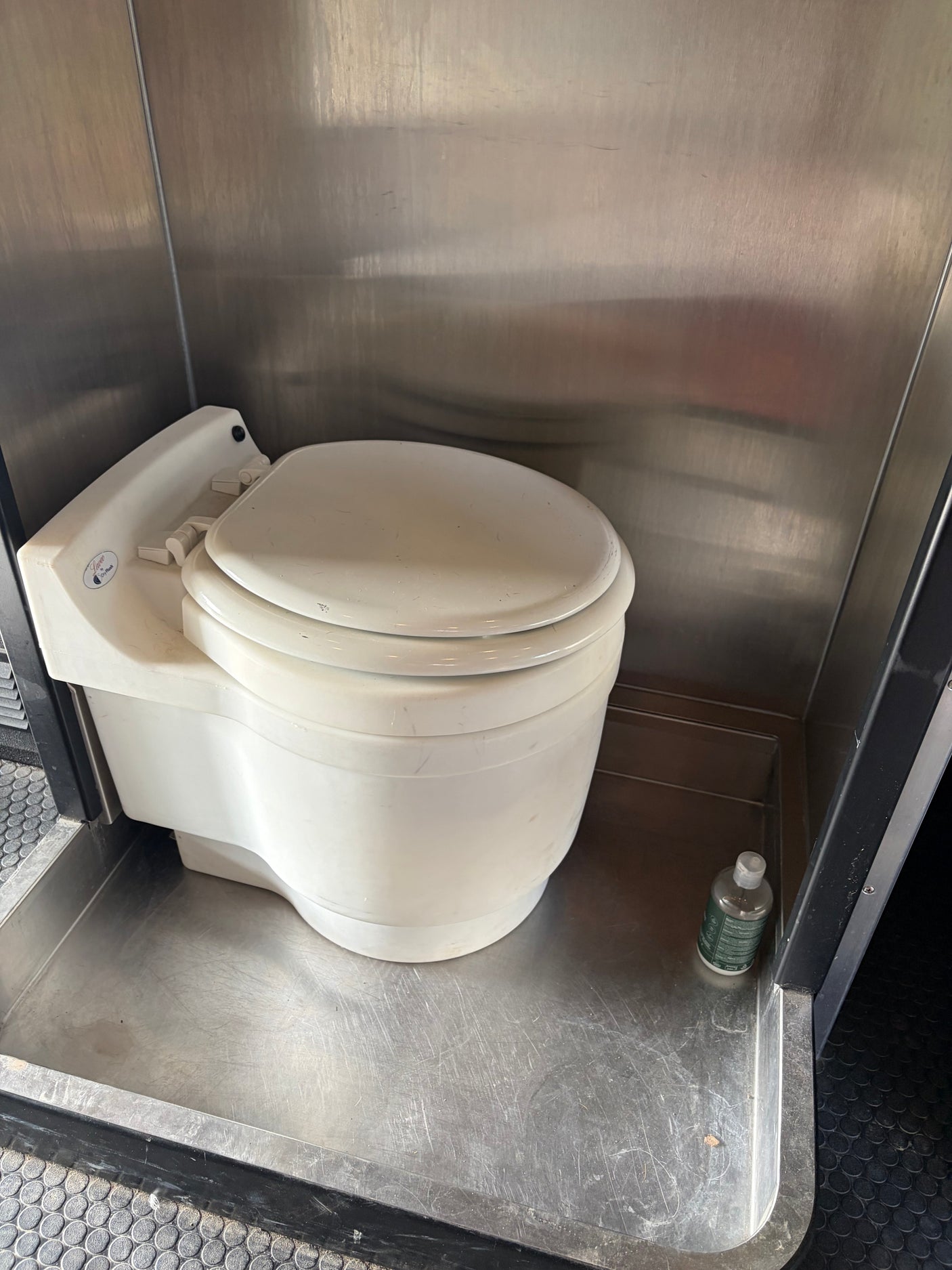 Laveo DryFlush composting toilet in an RV rental, featuring stainless steel walls and a small cleaning bottle on the floor.