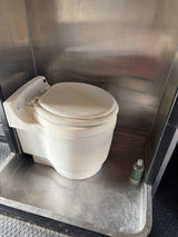 Laveo DryFlush composting toilet in an RV rental, featuring stainless steel walls and a small cleaning bottle on the floor.