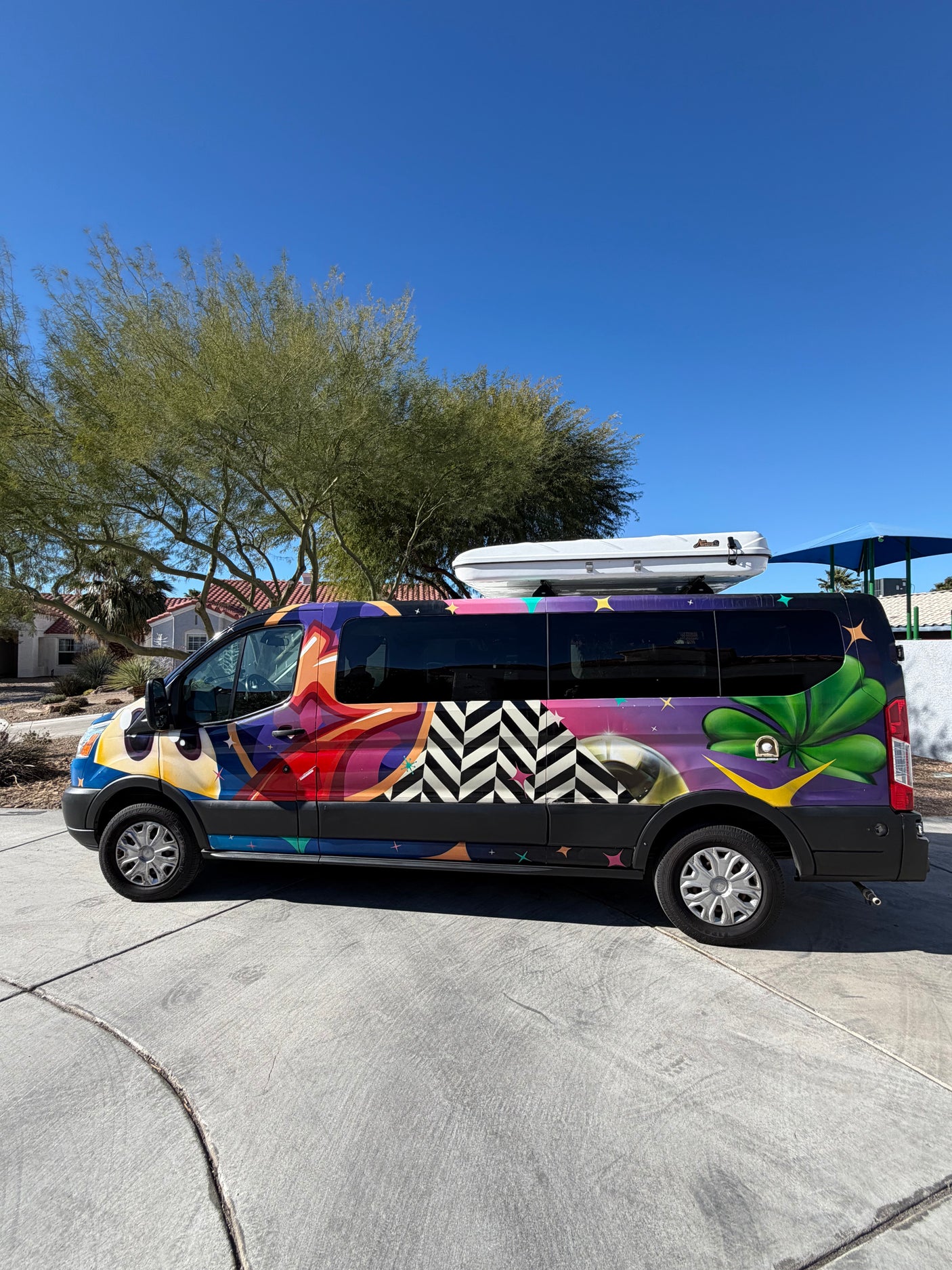 Colorful rv rental van, artistically wrapped, with a rooftop cargo box, parked outdoors on a sunny day.