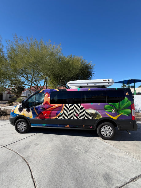 Colorful rv rental van, artistically wrapped, with a rooftop cargo box, parked outdoors on a sunny day.