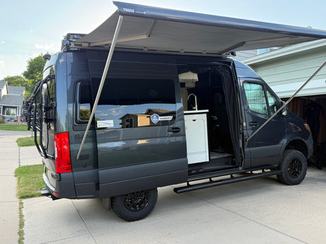 Dark grey camper van (RV rental) with open side door showing kitchen. Extended awning, roof AC, and rear bike rack visible.