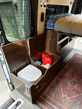 RV rental interior: Dometic portable toilet housed in a custom wooden enclosure, with a bed and integrated storage.