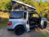 Adventure awaits! This grey Honda Element RV rental with a pop-top camper is ready for camping, doors open in a lush setting.