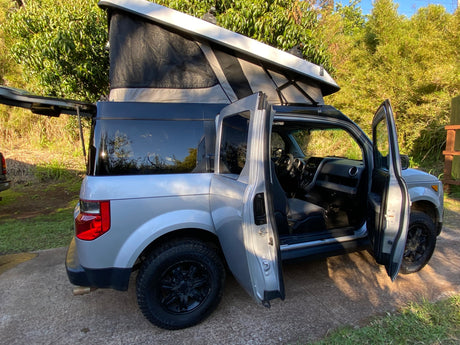 Adventure awaits! This grey Honda Element RV rental with a pop-top camper is ready for camping, doors open in a lush setting.