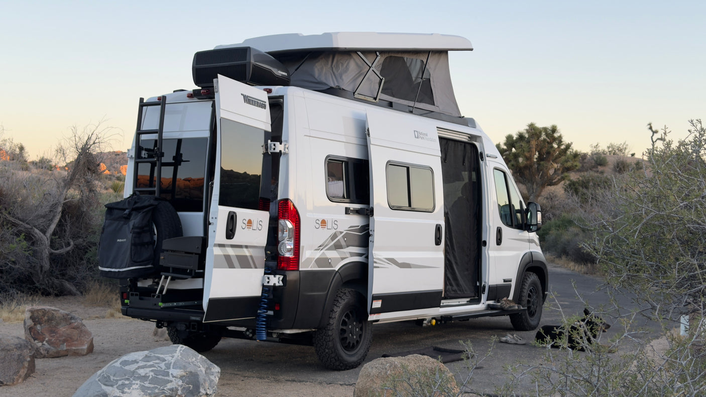 White Solis RV rental in a desert camp. Pop-top up, doors open, gear on back, and a dog enjoying the view.
