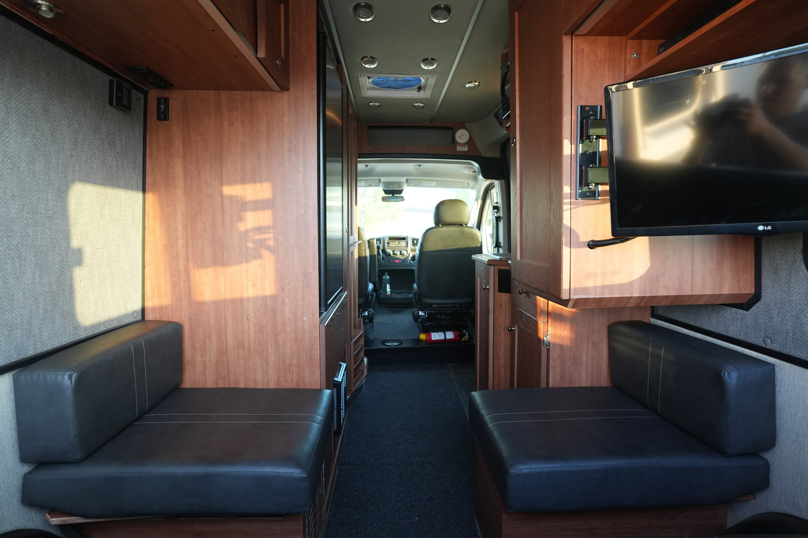 Interior of an rv rental: opposing dark benches, wood cabinetry, a TV, and the driver's cabin in view.