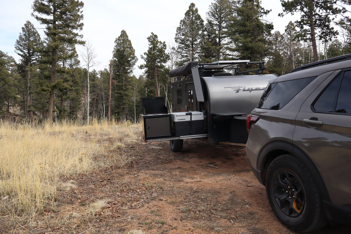 Adventure-ready rv rental: A dark SUV tows a Flyer camper trailer with an outdoor kitchen, set up in a pine-filled campsite.