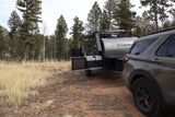 Adventure-ready rv rental: A dark SUV tows a Flyer camper trailer with an outdoor kitchen, set up in a pine-filled campsite.