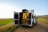 A white RV rental camper van with open rear doors on a dirt road, showcasing its inviting interior space & hill views.