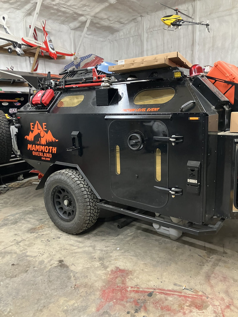 Side view of the 2023 Mammoth Overland ELE off-road trailer in a shop: side entry door with a round porthole window and two slit windows, orange MAMMOTH OVERLAND ELE graphics on the black body panel, roof rack loaded with recovery gear and a fuel can, and an all-terrain tire on a black wheel — photo 2