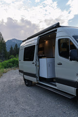 Light grey RV rental camper van parked on a gravel road with open side door, showing interior. Mountains and trees in background.
