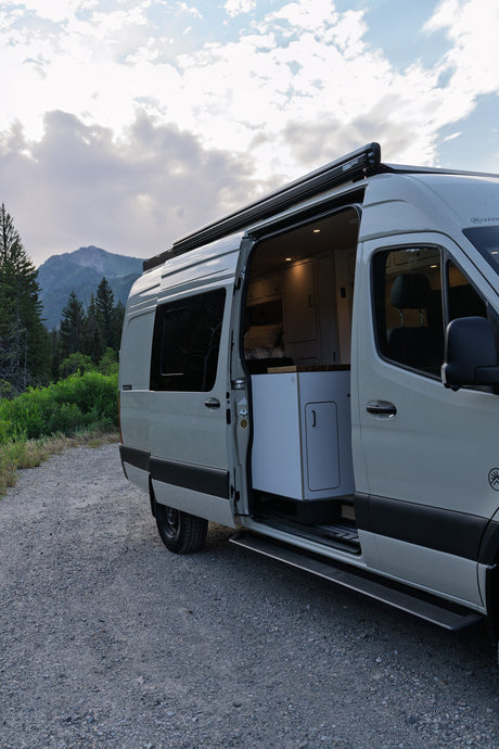 Light grey RV rental camper van parked on a gravel road with open side door, showing interior. Mountains and trees in background.