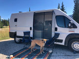 A dog stands on a colorful mat beside a white rv rental camper van with its side door open, chairs set up outside.