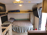 This RV rental interior features a cozy bed, kitchenette, TV, and an upper bunk, perfect for compact travel.