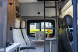 Inside a modern RV rental: comfortable quilted seats, a workstation with a laptop & sunflower, and a ladder to an overhead bed.