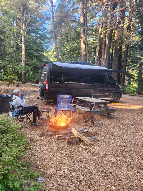 Person enjoying a campfire at a wooded RV rental campsite with a black van, picnic table, and chairs.