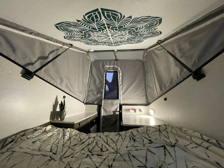 Inside a cozy RV rental: a patterned bed, mandala ceiling art, and gray pop-up fabric walls.