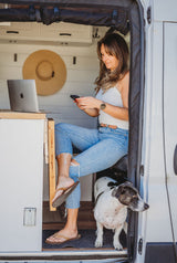 Woman sits in an RV rental with phone & laptop, dog nearby. Van life dream for remote workers.