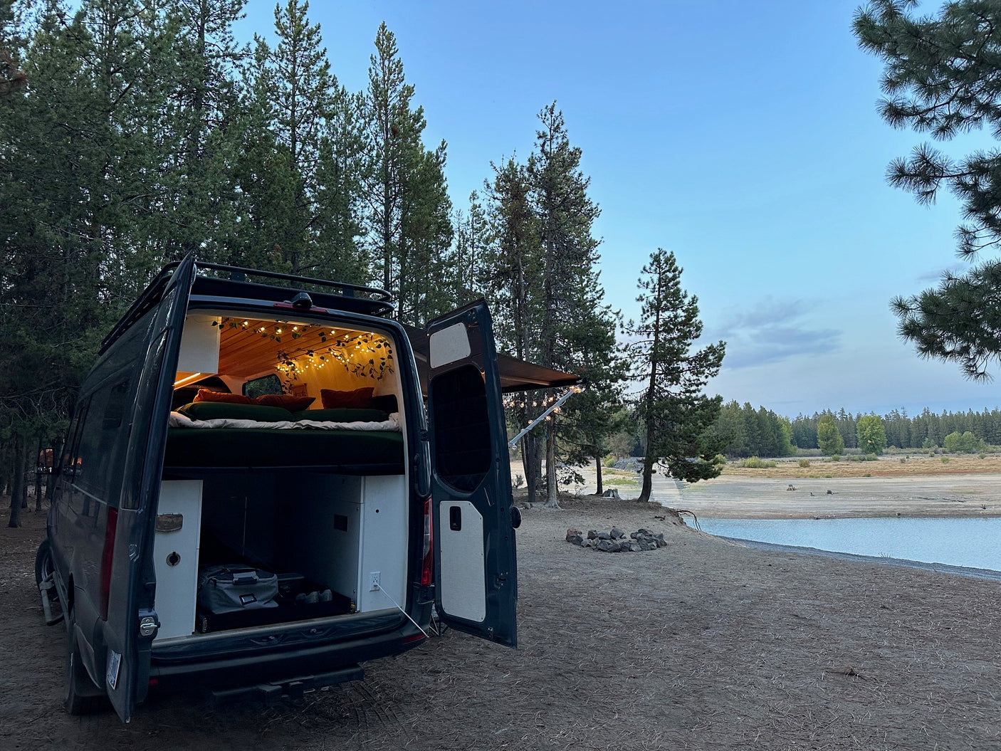 Experience van life: an RV rental with illuminated interior & comfy bed is parked on a sandy shore next to a lake.
