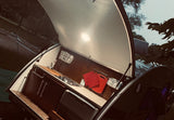 Compact kitchen of an RV rental teardrop camper with open hatch and light. Features sink, stove, cabinets, cooler, & red pot holders. Ready for travel!