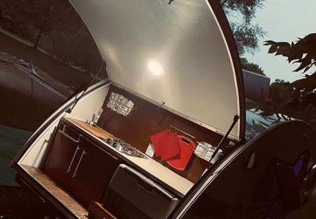 Compact kitchen of an RV rental teardrop camper with open hatch and light. Features sink, stove, cabinets, cooler, & red pot holders. Ready for travel!