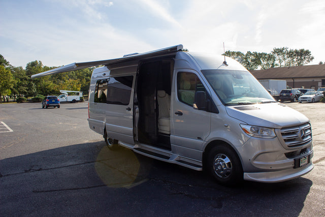 This luxury silver Mercedes-Benz Sprinter rv rental features an extended awning and open door, revealing its interior.