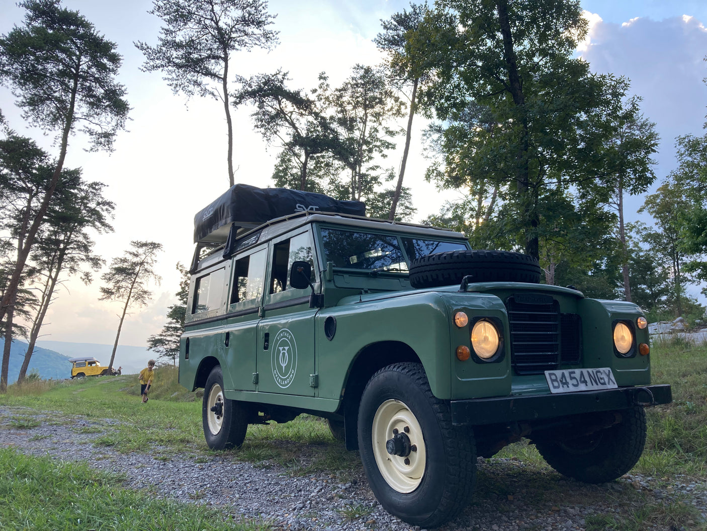 Green vintage Land Rover with rooftop tent, ideal for an RV rental overlanding trip. A child and another vehicle are visible.