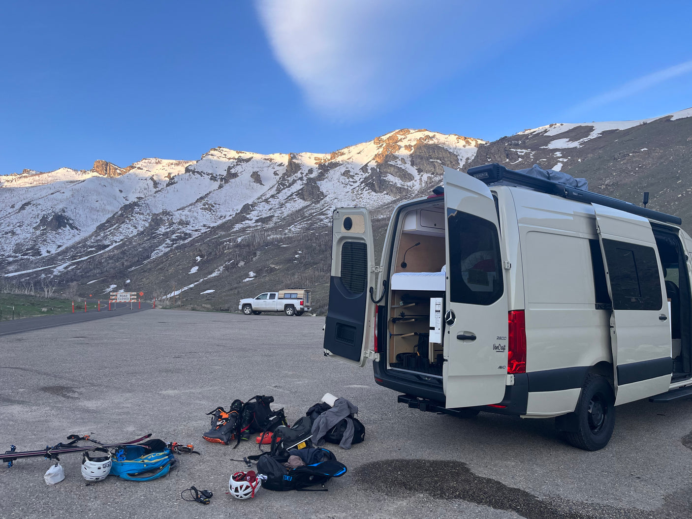 White rv rental van with open doors displaying interior bed/storage, parked in snowy mountains with ski/snowboard gear on the ground.