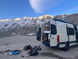 White rv rental van with open doors displaying interior bed/storage, parked in snowy mountains with ski/snowboard gear on the ground.
