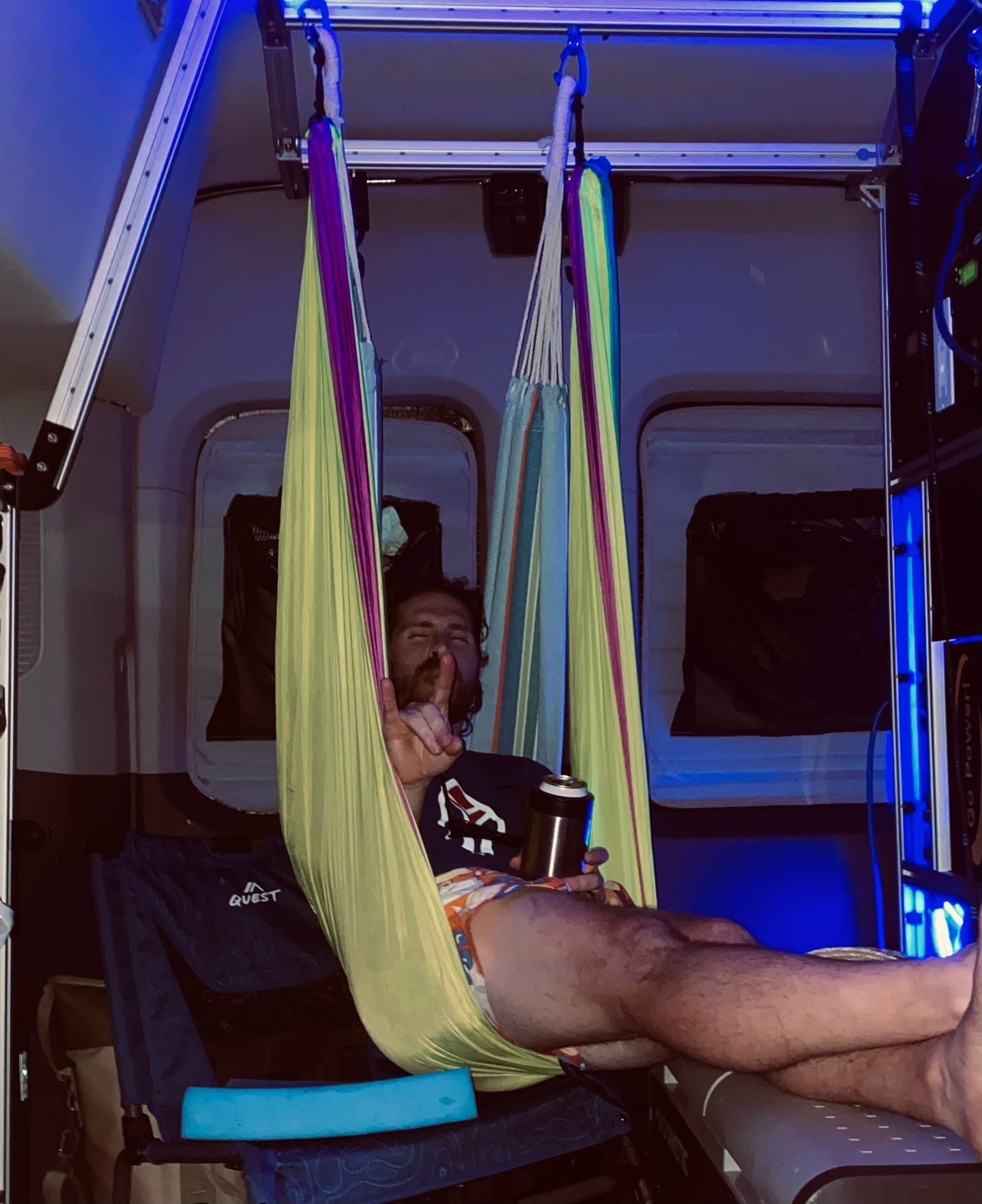 Inside an RV rental camper van, a man relaxes in a colorful hammock under blue lights, holding a drink.