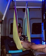 Inside an RV rental camper van, a man relaxes in a colorful hammock under blue lights, holding a drink.