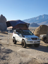 Lexus GX with rooftop tent & ladder parked among desert boulders. Great off-road RV rental alternative for mountain adventures. Snow peaks visible.