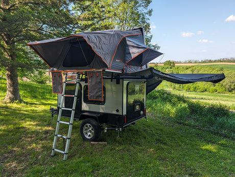 Compact rv rental trailer with a rooftop tent and awning, set up for a comfortable camping adventure in a scenic landscape.