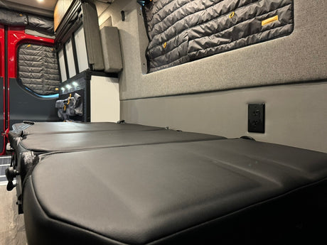 Explore this RV rental's cozy interior: modular black seating/bed, power outlet, insulated windows, ideal for mobile adventures.
