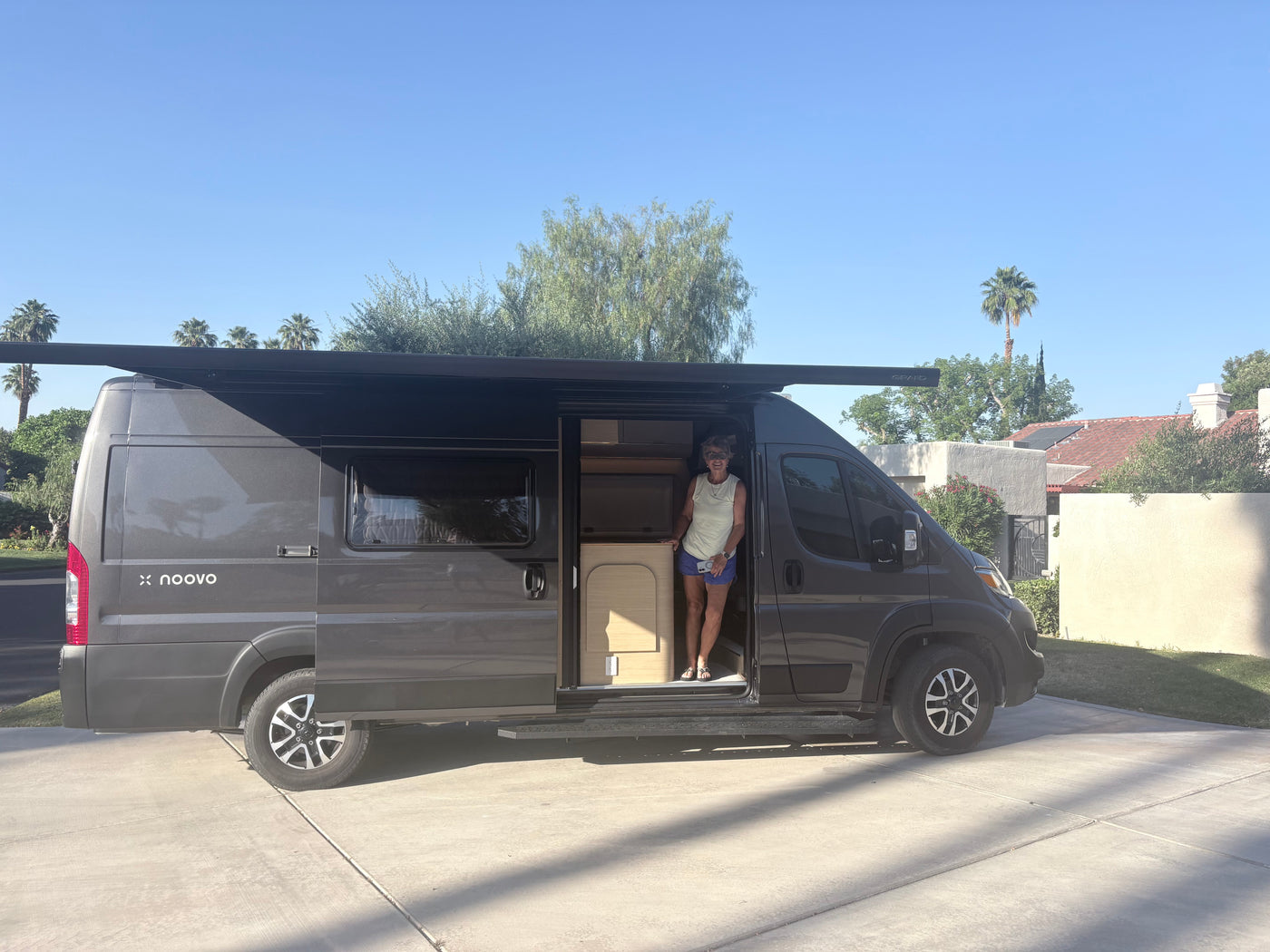 A woman smiles in her dark grey Noovo rv rental van with an extended awning. Ready for travel under sunny skies. — photo 2