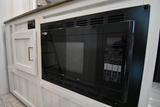 Built-in black microwave and light wood cabinetry in an RV rental kitchen, showing an electrical outlet and cabinet door.