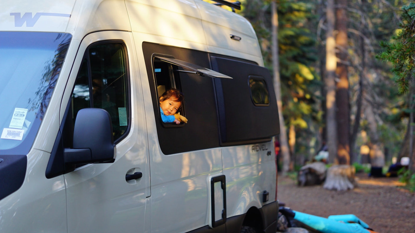 A joyful child peeks from a light-colored RV rental window, parked amidst sunlit trees. Ideal for family camping trips.