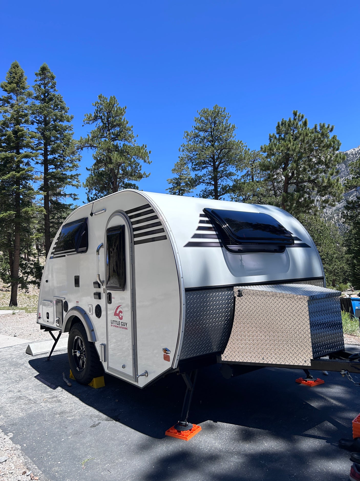 White teardrop RV rental parked amidst pine trees under a clear blue sky, featuring black and white striped accents. Ideal for camping.