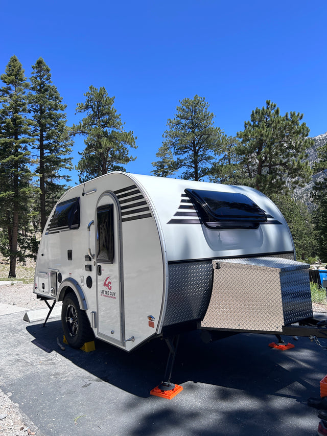 White teardrop RV rental parked amidst pine trees under a clear blue sky, featuring black and white striped accents. Ideal for camping.