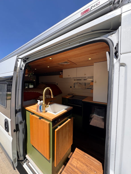 Open the door to this inviting rv rental camper van. View of a modern kitchen with sink, golden faucet, wooden ceiling, and bed.