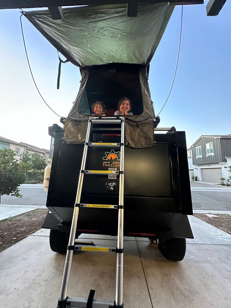Two smiling girls look from an Off Grid Trailer rooftop tent, a unique RV rental experience. The trailer and ladder are visible, ready for adventure!