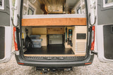 Rear of a custom camper van showing organized under-bed storage with a fridge, vacuum, and cables. Great for an RV rental.