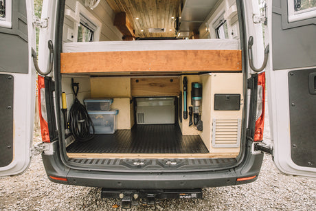 Rear of a custom camper van showing organized under-bed storage with a fridge, vacuum, and cables. Great for an RV rental.
