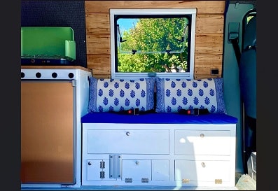 Cozy RV rental interior featuring a compact camper kitchen, comfortable blue seating with storage, and a scenic window. Perfect for van life. — photo 10
