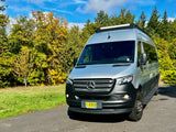 A sleek silver & black Mercedes-Benz Sprinter rv rental, featuring a roof light bar, parked on asphalt with autumn trees and blue sky.