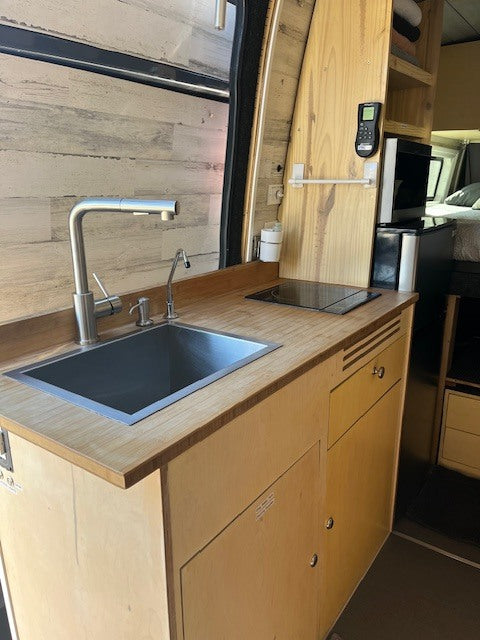 RV rental interior: A bright camper van kitchen with wood counters, sink, induction cooktop, microwave, and fridge.