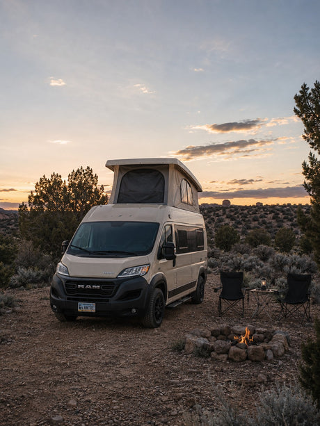 Enjoy a desert sunset from your cozy RV rental, featuring a tan camper van, campfire, and seating for two.