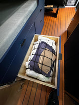 RV rental storage: A deep, open drawer under a dark blue bunk bed, holding bedding secured by a net. Efficient camper organization.