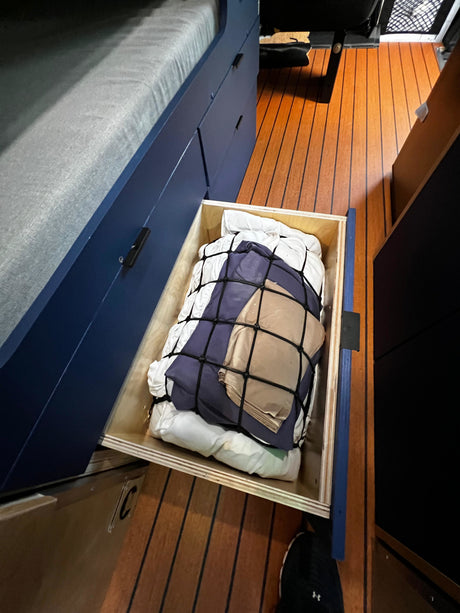 RV rental storage: A deep, open drawer under a dark blue bunk bed, holding bedding secured by a net. Efficient camper organization.